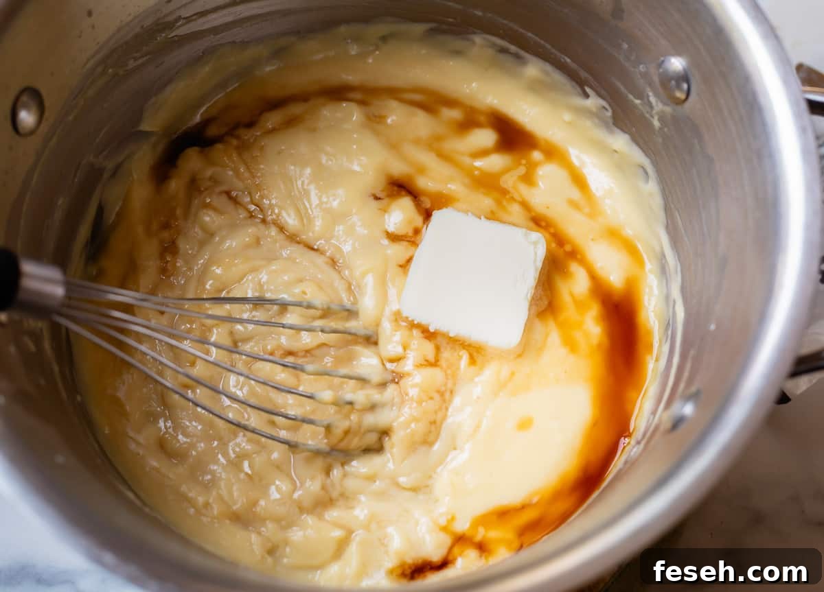 butter and vanilla being stirred into thickened pastry cream in a silver pot.