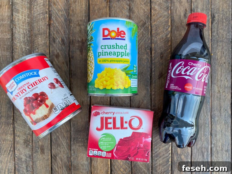 Cherry Coke Jello Salad Recipe — Retro Fizzy Dessert 5 Ingredients to make cherry jello coke salad are a can of cherry pie filling, crushed pineapple, cherry jello and cherry coca cola on a wooden counter top.