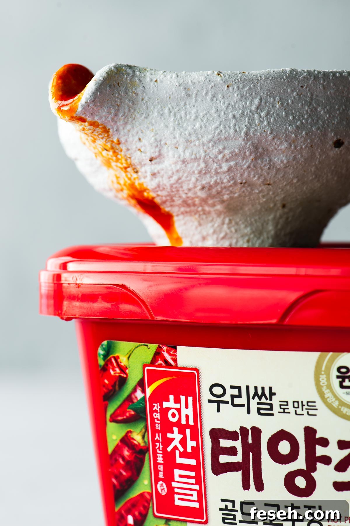 Side angle view of a pourer with red bibimbap sauce drizzling from the spout, on top of a red tub of gochujang paste.