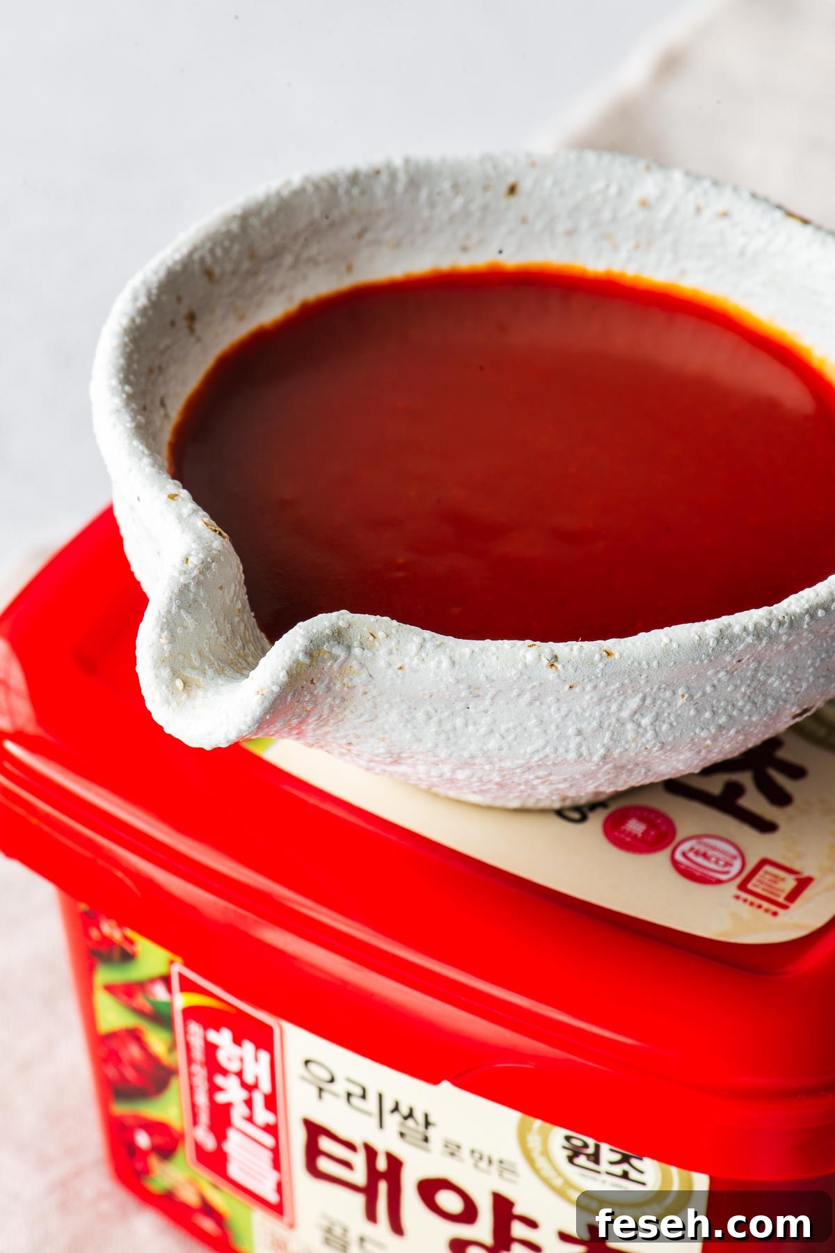 Close-up of bibimbap sauce in a white ceramic cup on top of a tub of gochujang paste.