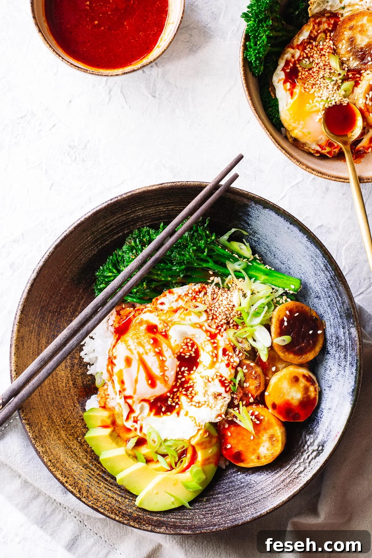 Top down view of a Korean rice bowl (bibimbap) with sweet potato, broccoli, avocado, a fried egg and gochujang sauce, with chopsticks and sauce bowl..