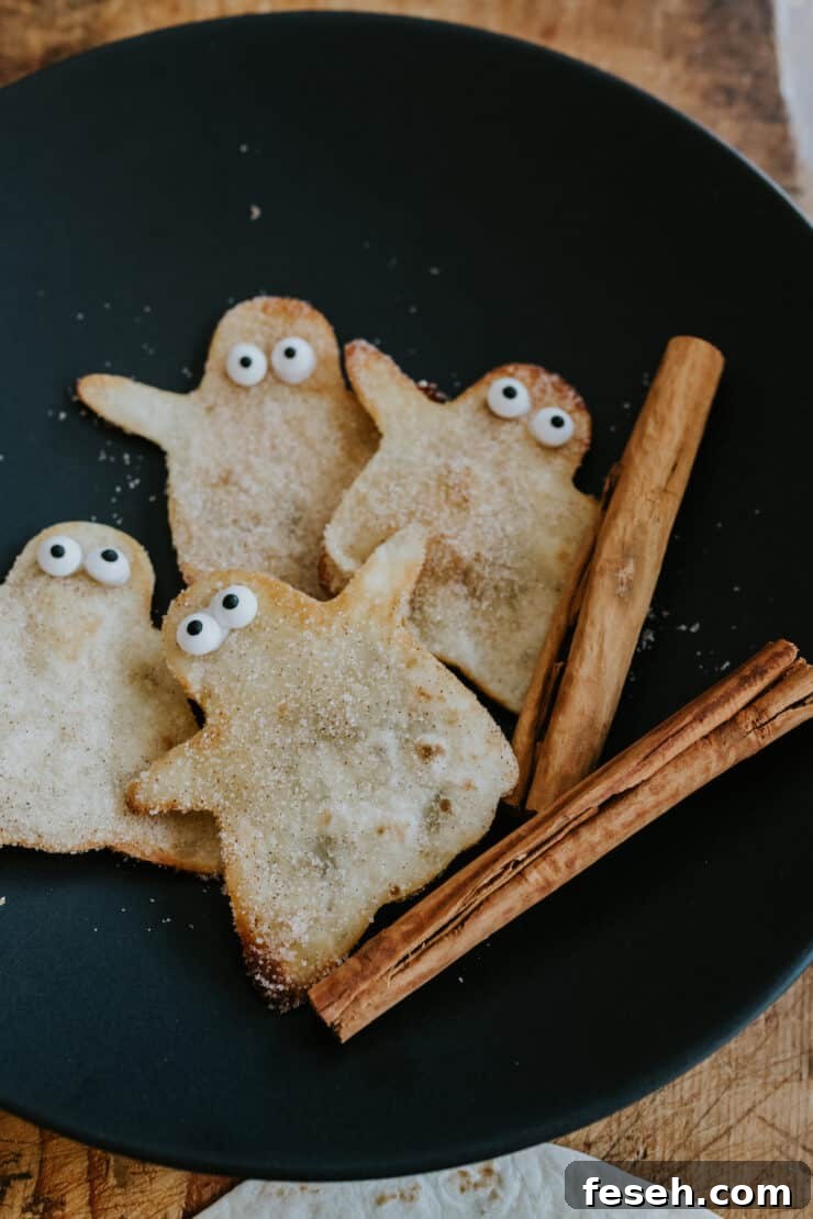 Cinnamon-sugar coated fried buñuelo ghosts on a black plate with cinnamon sticks on the side.