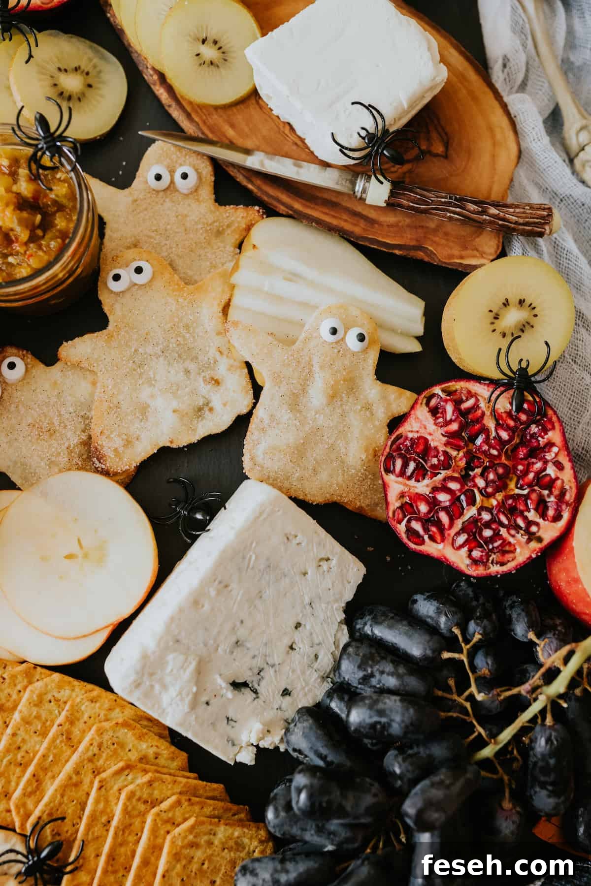 Close-up of cinnamon-sugar buñuelo ghosts with candy eyeballs surrounded by cheese and fresh fruit on a Halloween cheese board.