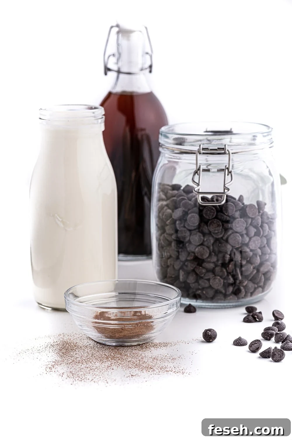 Jars filled with fresh whipping cream, chocolate chips, expresso powder and vanilla sitting on a bright white table against a bright white background. 