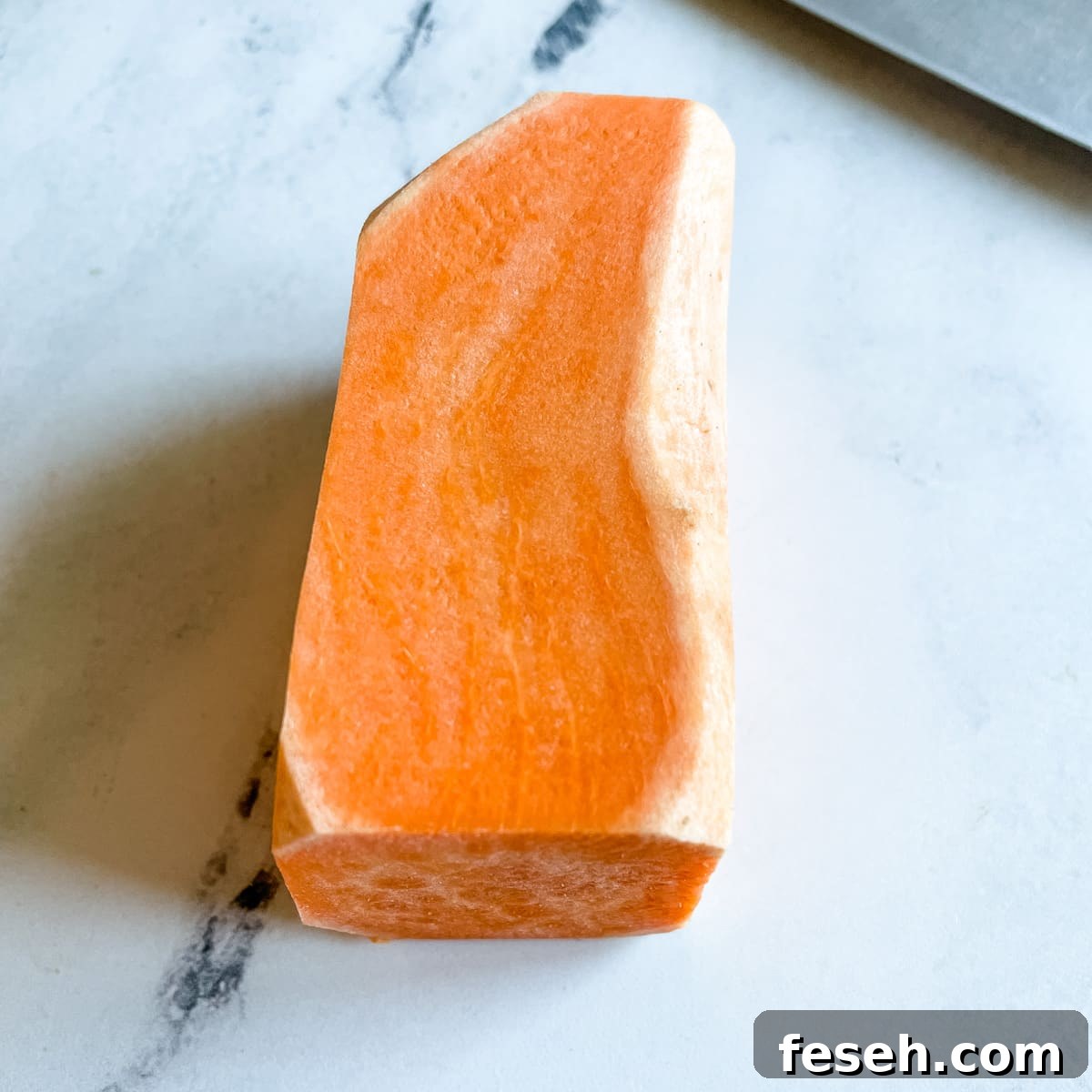 A peeled sweet potato with the round sides trimmed to create a rectangle sits on a white marble counter top next to the blade of a kitchen knife.