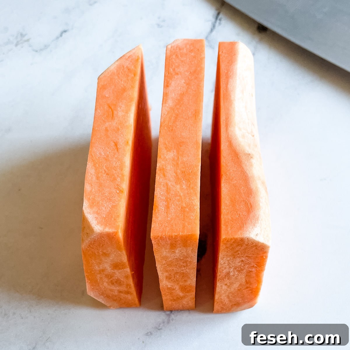 A peeled sweet potato cut horizontally into 3 equal pieces sits on a white marble counter top next to the blade of a kitchen knife