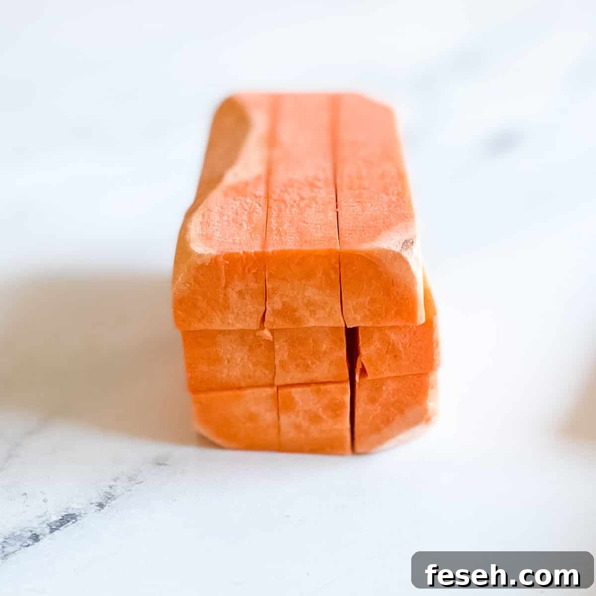 A peeled sweet potato cut both horizontally and diagonally into equal pieces to create long sticks sits on a white marble counter top.