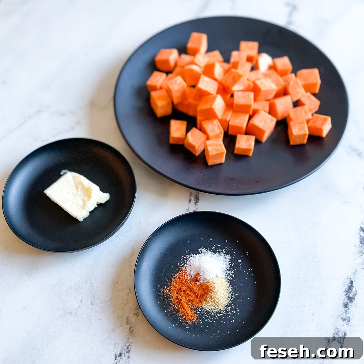 A large black plate with cubed sweet potatoes, a small black plate with a pat of butter, and a small black plate with cayenne, garlic powder, and salt sit on a white marble counter top