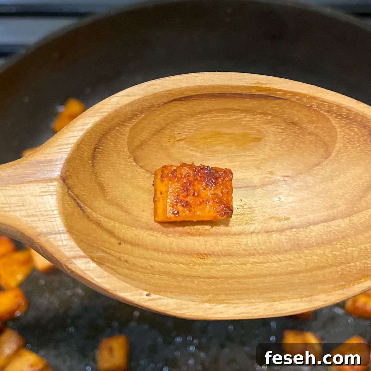 A wooden spoon in the foreground shows a browned cube of sweet potato over a black frying pan with sweet potato cubes cooking in it.