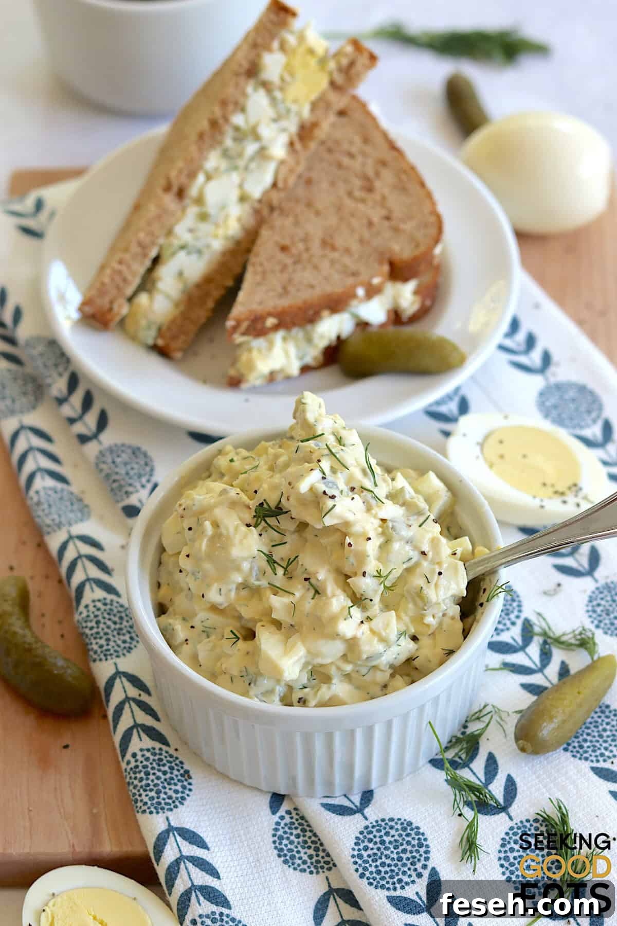Egg salad with dill pickles in small white ramekin and a silver spoon, next to an egg salad sandwich on a small round white plate.