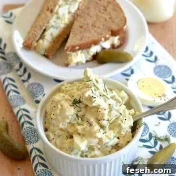 Egg salad with dill pickles served in a small white ramekin and a silver spoon, next to an egg salad sandwich on a small round white plate.