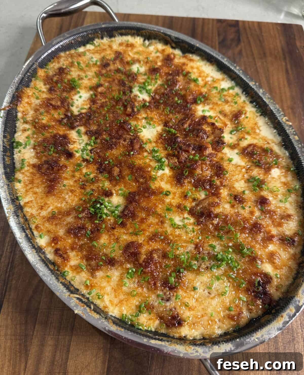 A baked dish with a golden-brown, crispy cheese and breadcrumb topping, sprinkled with finely chopped green herbs, sits in an oval metal pan on a wooden surface.