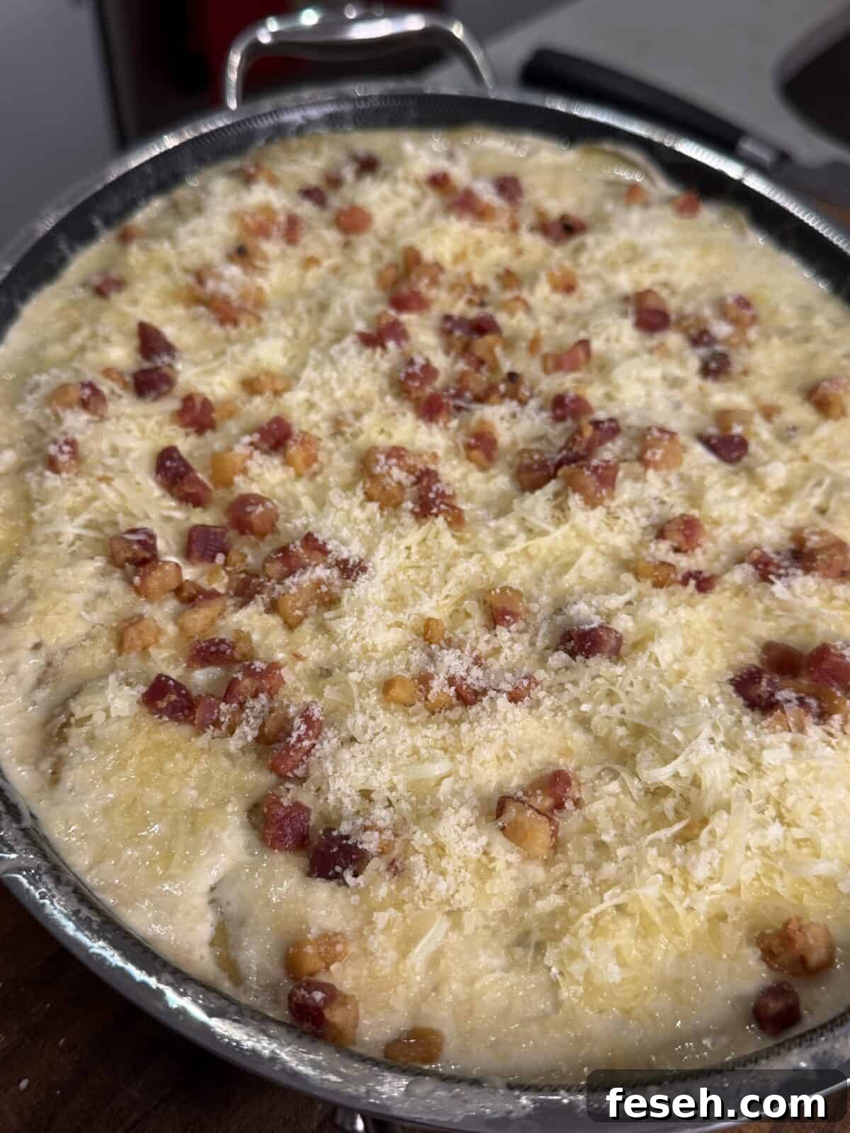 A close-up of an uncooked dish topped with grated cheese and small cubes of pancetta or bacon, in a metal baking pan, ready to be baked.