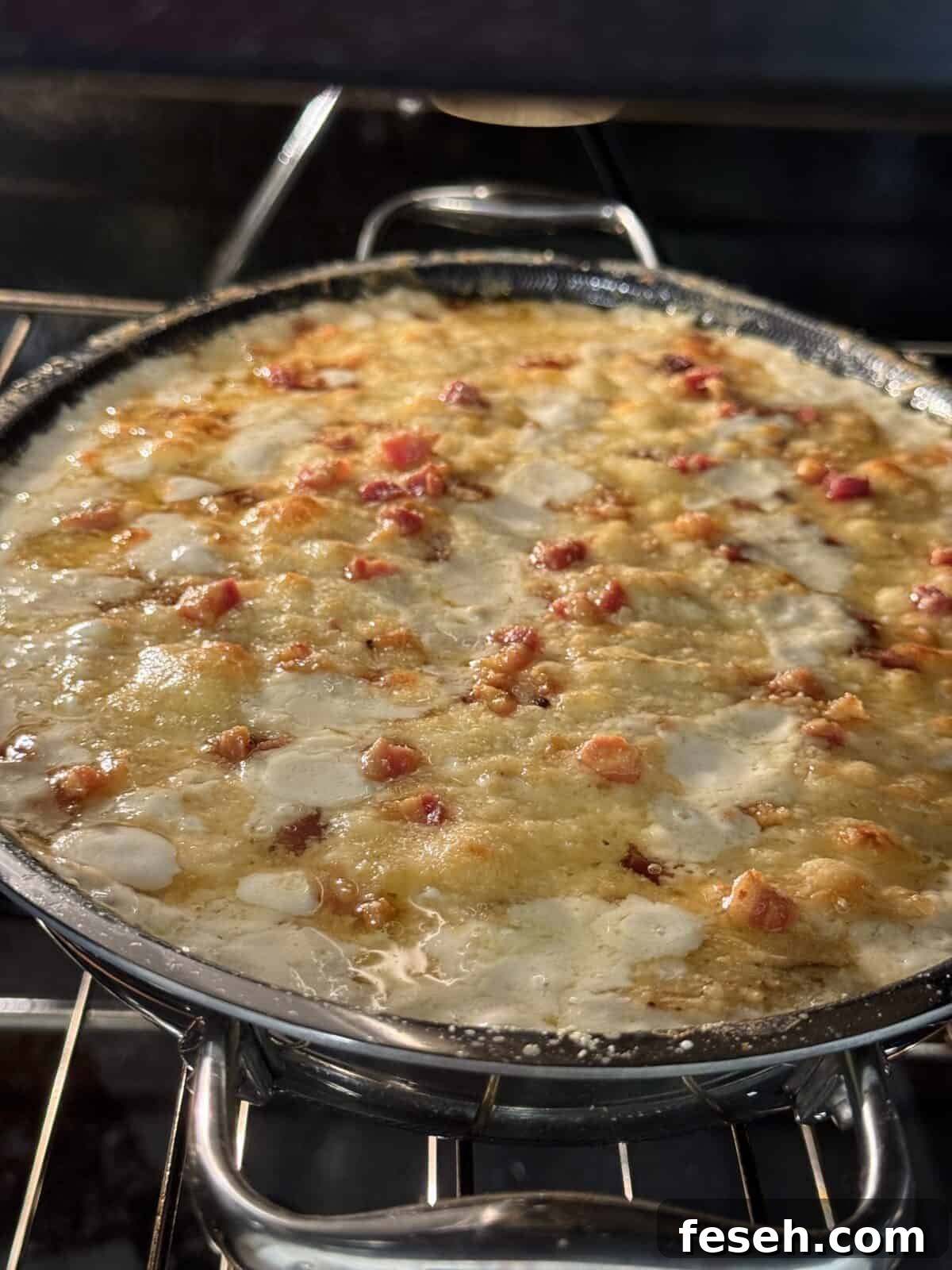 A close-up of a cheesy dish with bubbling melted cheese and pieces of bacon or ham, baking in a round pan inside an oven. The surface appears golden and slightly crispy.