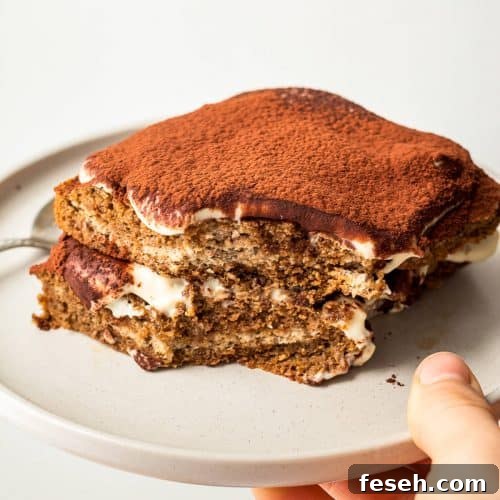 A hand holding a grey plate with a single serve tiramisu with a bite missing.