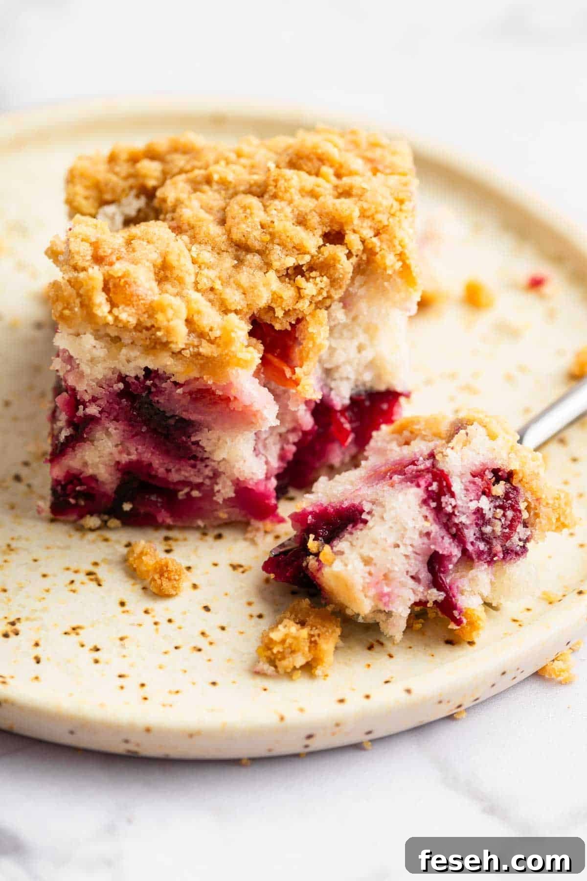 Square slice of plum cake on plate with fork removing a bite.