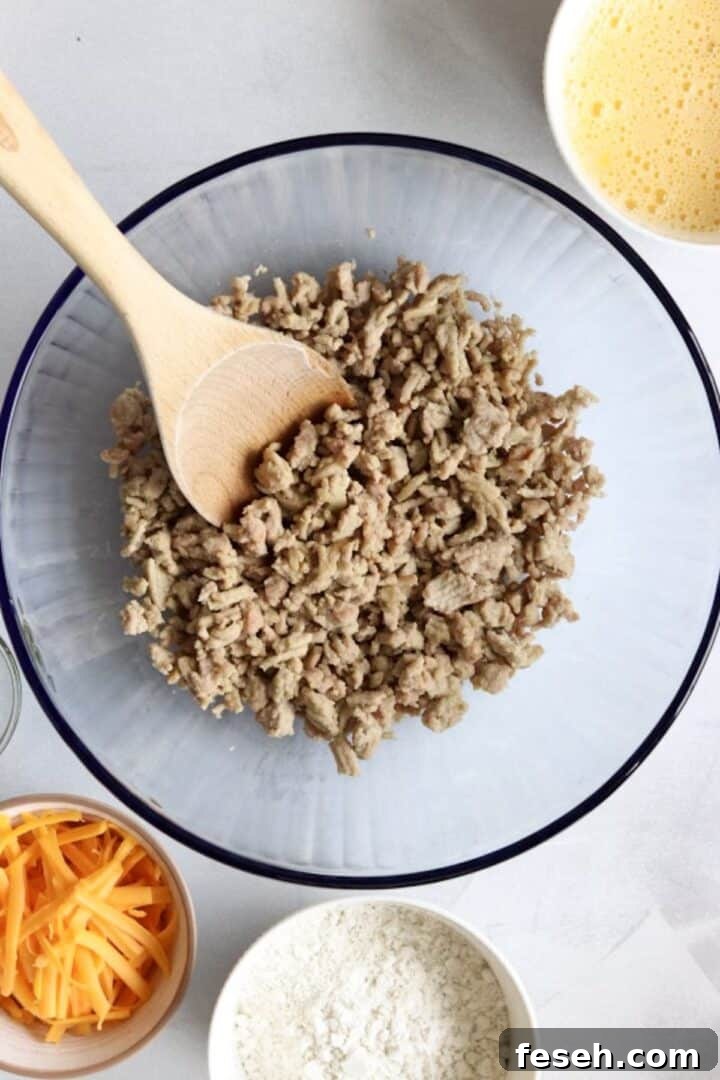 Cooked and crumbled chicken sausage in a large glass bowl with a wooden spoon