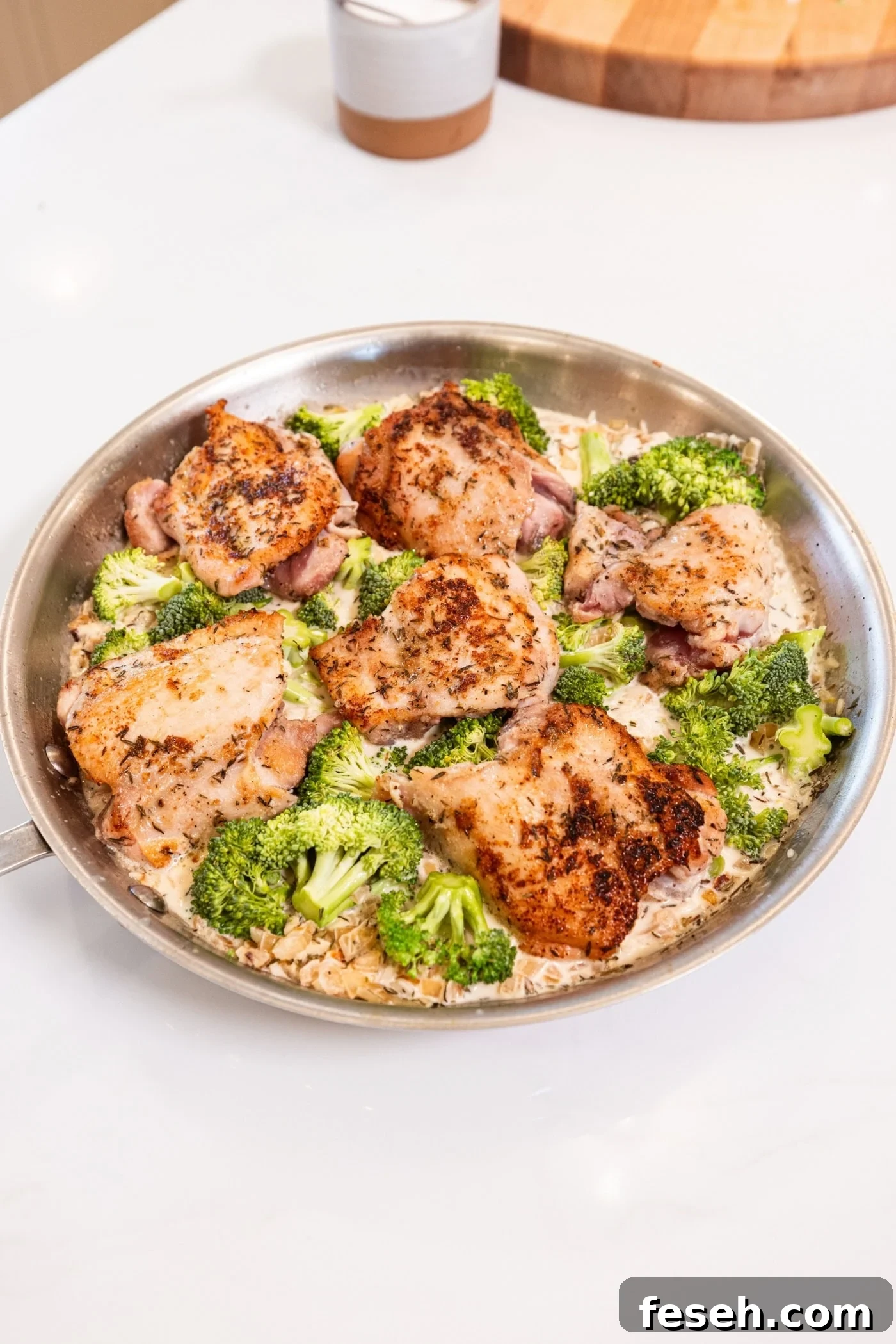 Metal skillet on a white surface filled with partially cooked chicken thighs and rice and broccoli.