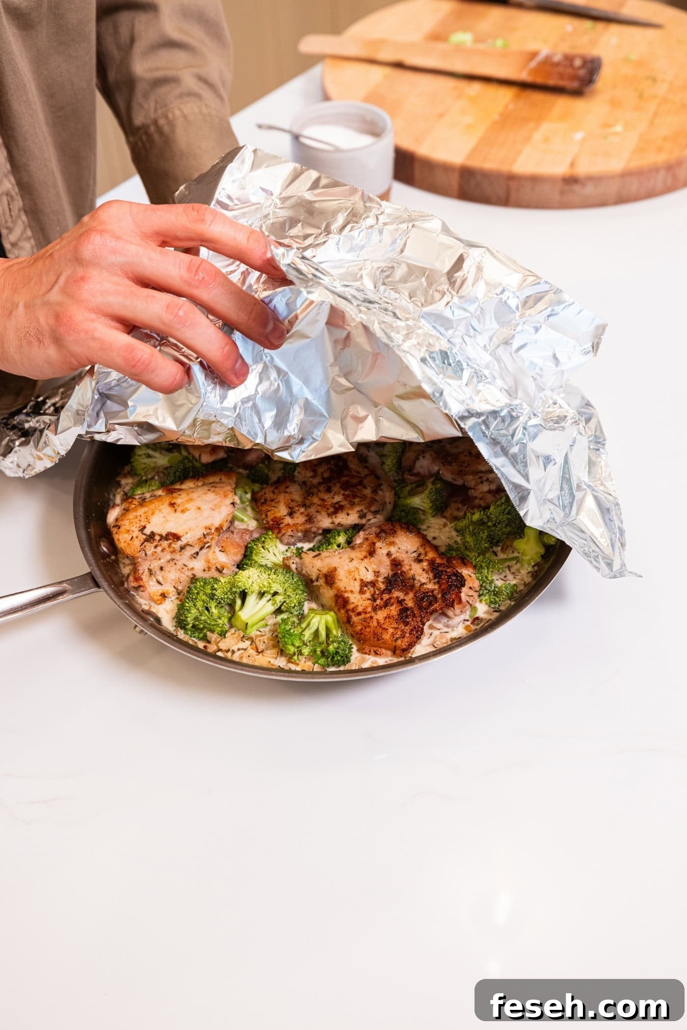 Hand pulling back cover on skillet filled with cooking chicken, broccoli, and rice.