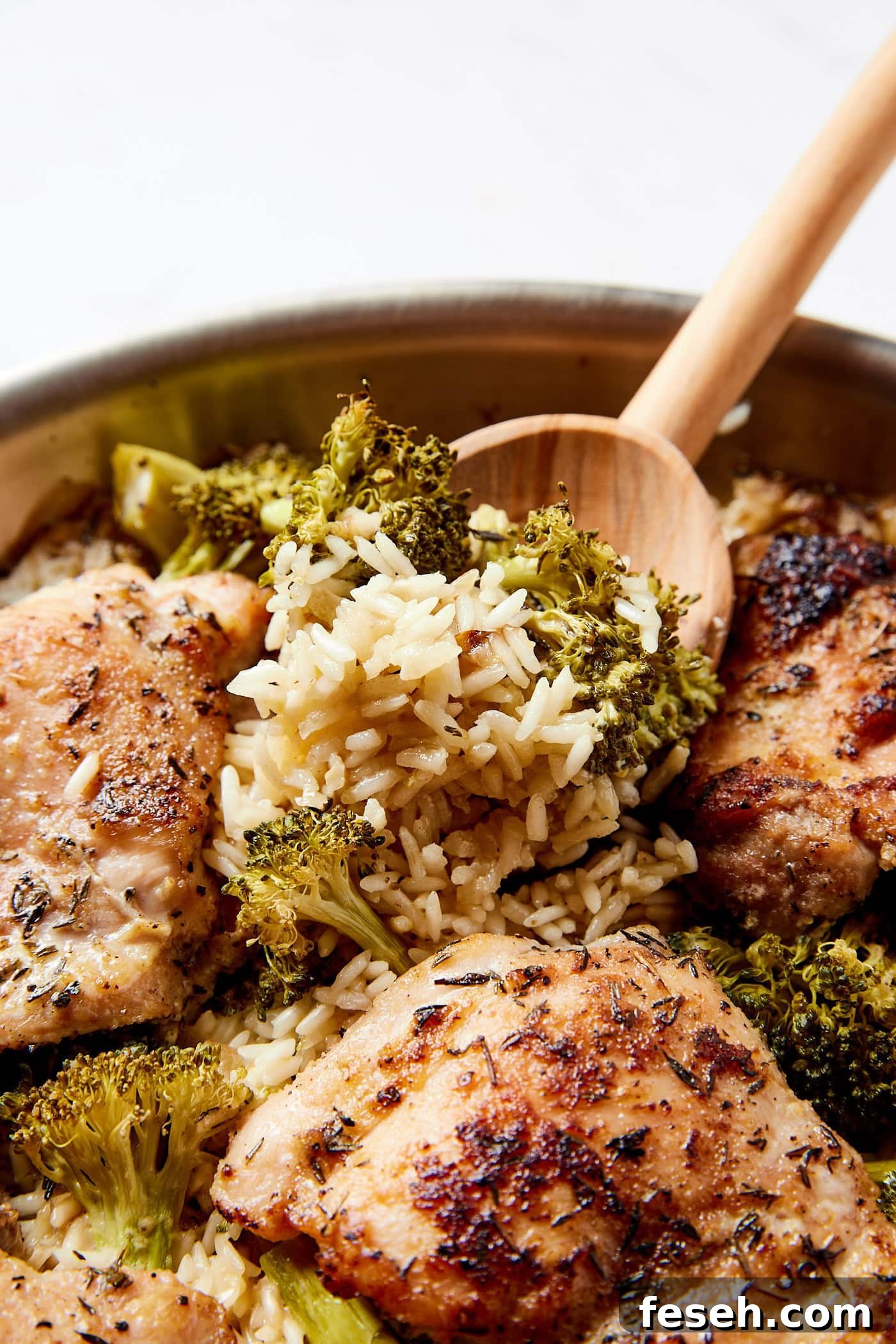 Wooden spoon scooping up a serving of rice with broccoli from a metal skillet filled with chicken thighs.