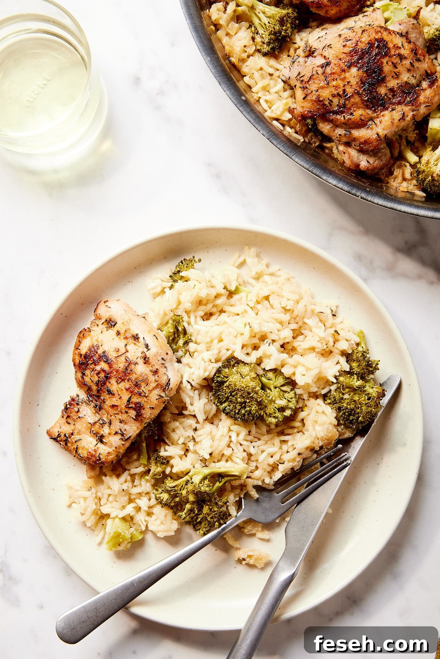 White marble surface with plate with serving of chicken and rice and broccoli with utensils resting on the plate.