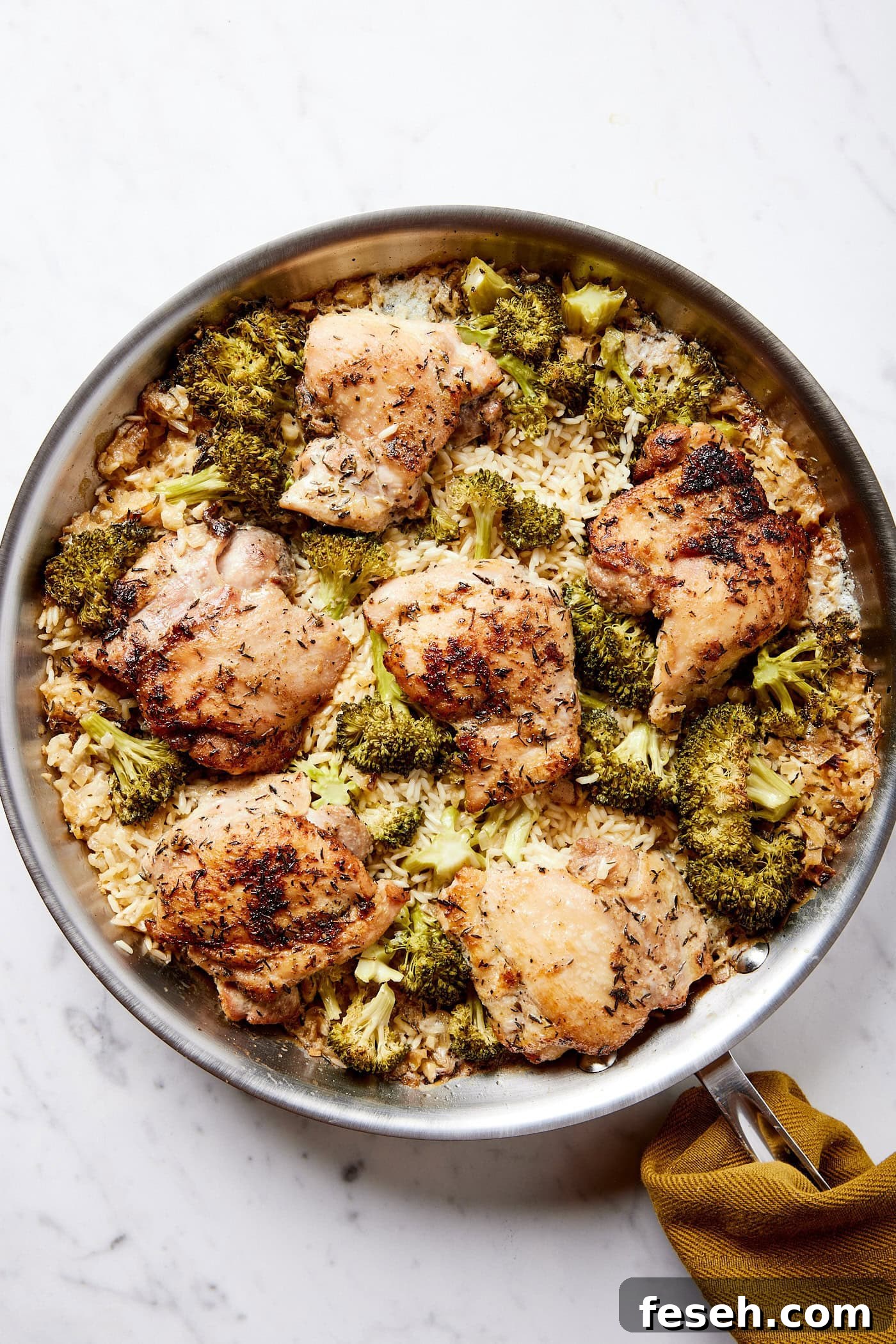 Metal skillet on white marble surface filled with cooked chicken, broccoli, and rice casserole with browned chicken texture.