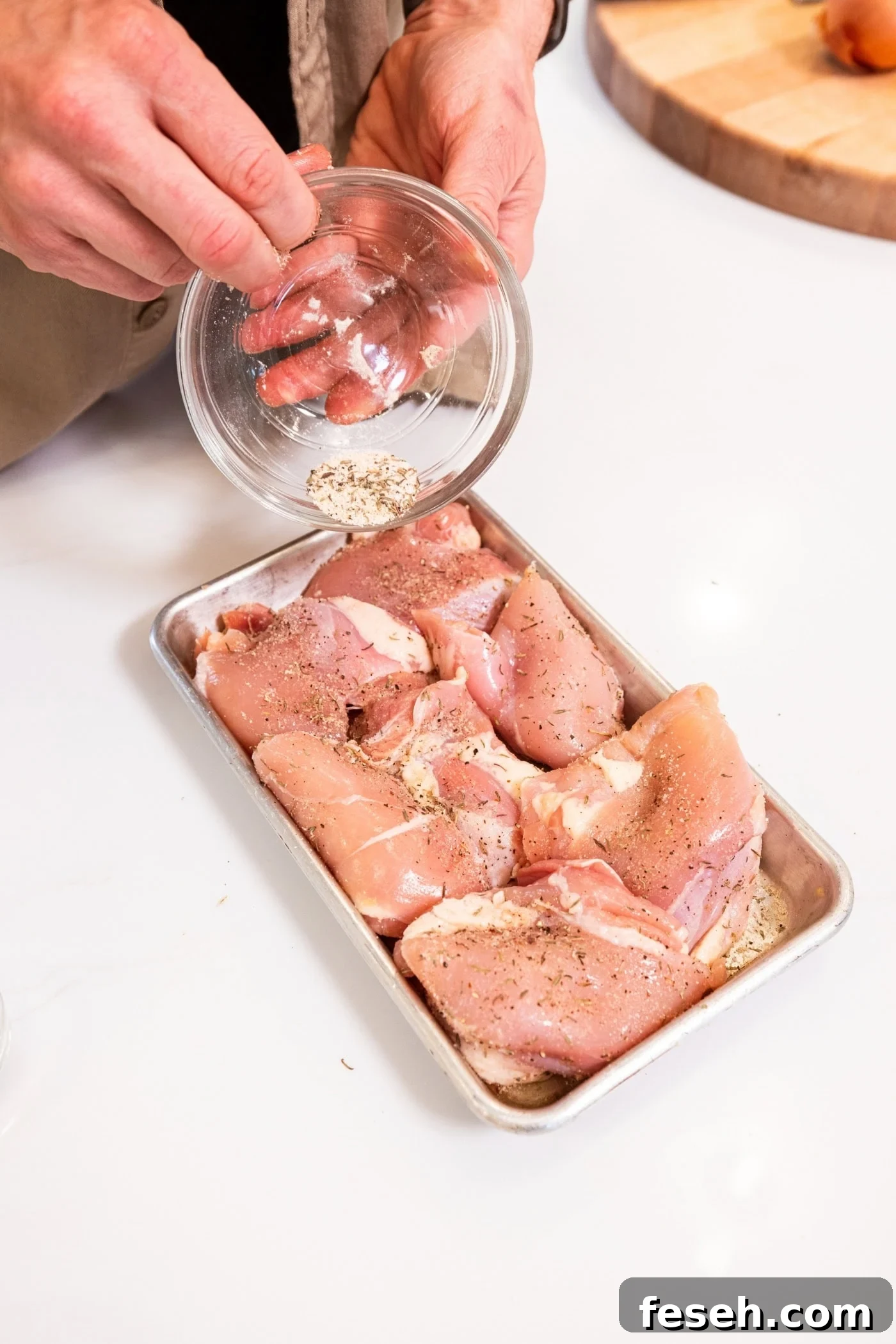 Hand pouring dry rub on top of raw chicken thighs on a small metal sheet before letting marinate.