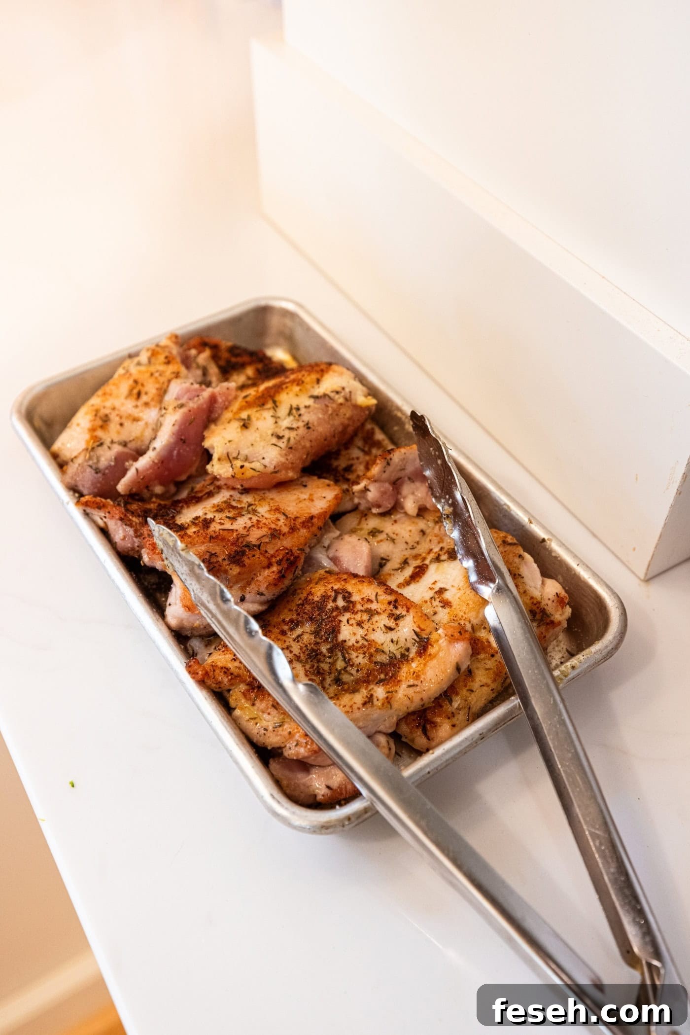Chicken thighs after being browned on one side sitting on a metal pan with tongs.