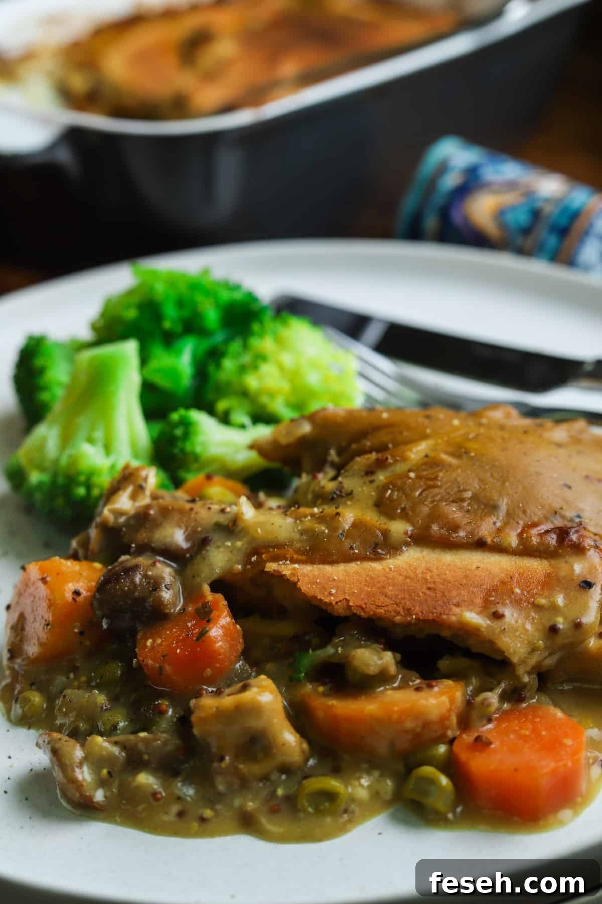 Close-up photo of a serving of vegan pot pie with broccoli on a white plate with cutlery and the main dish in the background.
