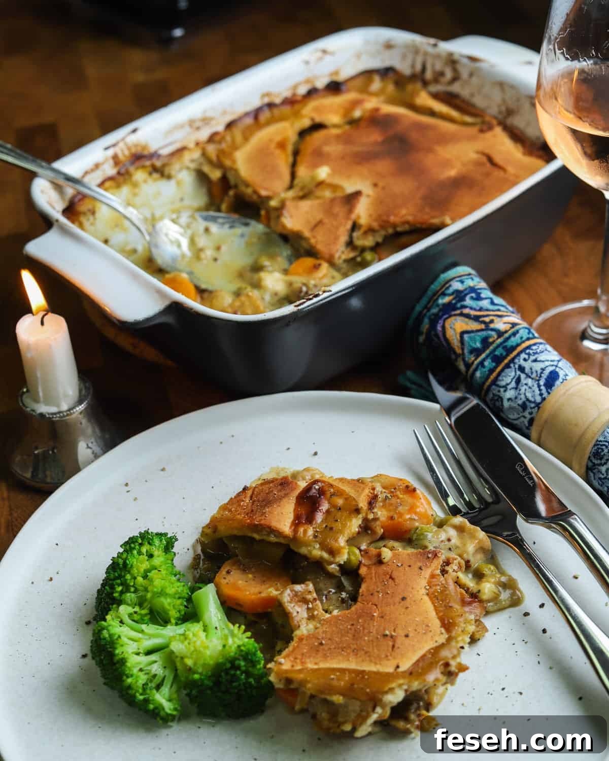 Photo of a plate of vegan pot pie with broccoli and the serving dish in the background and a candle, glass of wine and napkin.