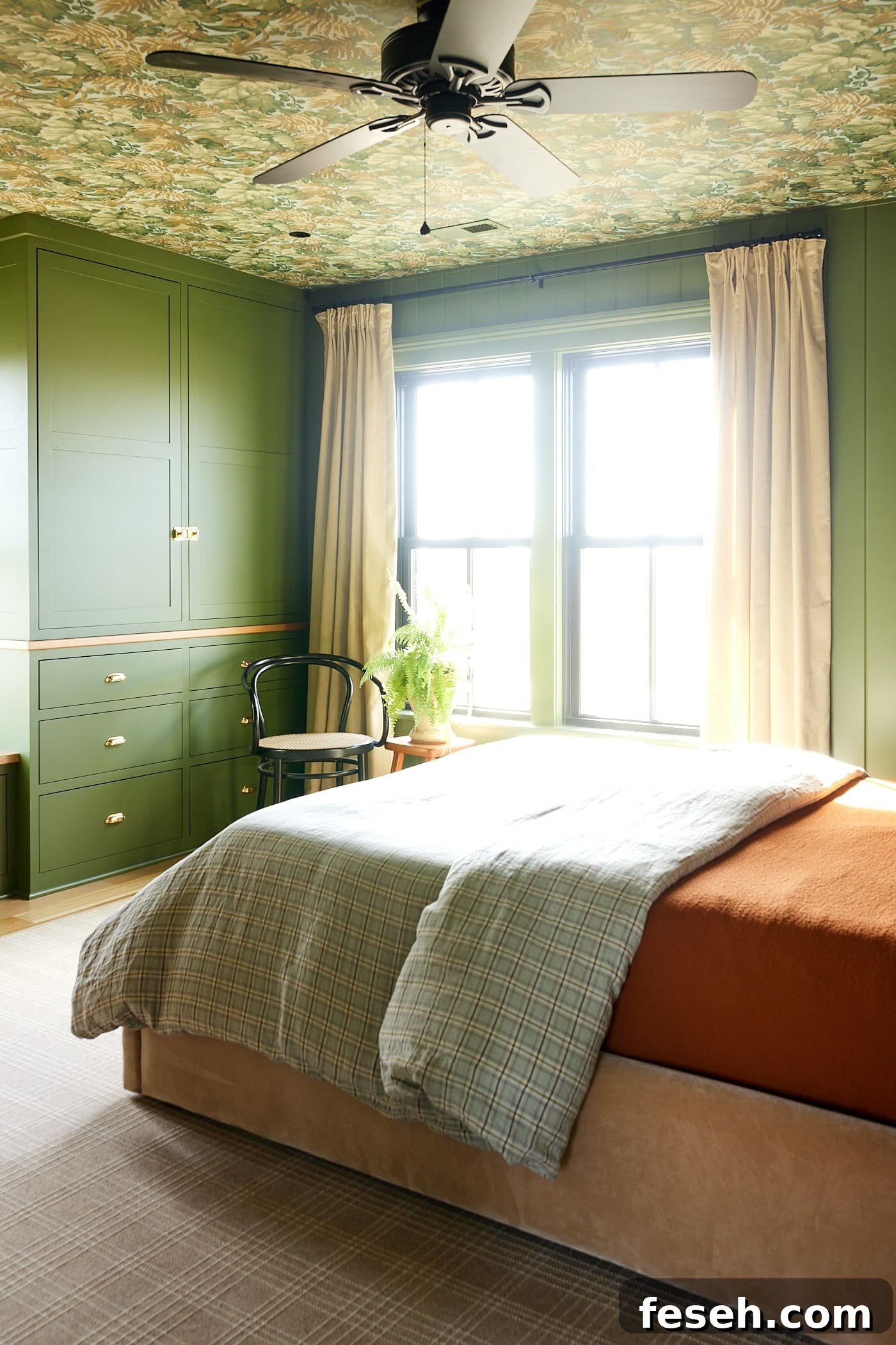 Green colored bedroom with large windows and built-in cabinets to the side with queen-sized bed with orange comforter and wallpaper on the ceiling.