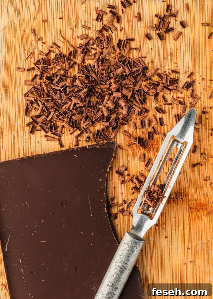 A bar of chocolate, vegetable peeler, and chocolate shavings on a wood board.