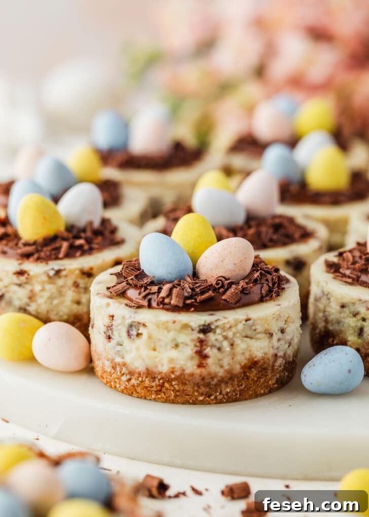 A white platter of mini Easter egg cheesecakes on a tan counter next to pink flowers, a brown bowl of candies, and white eggs.