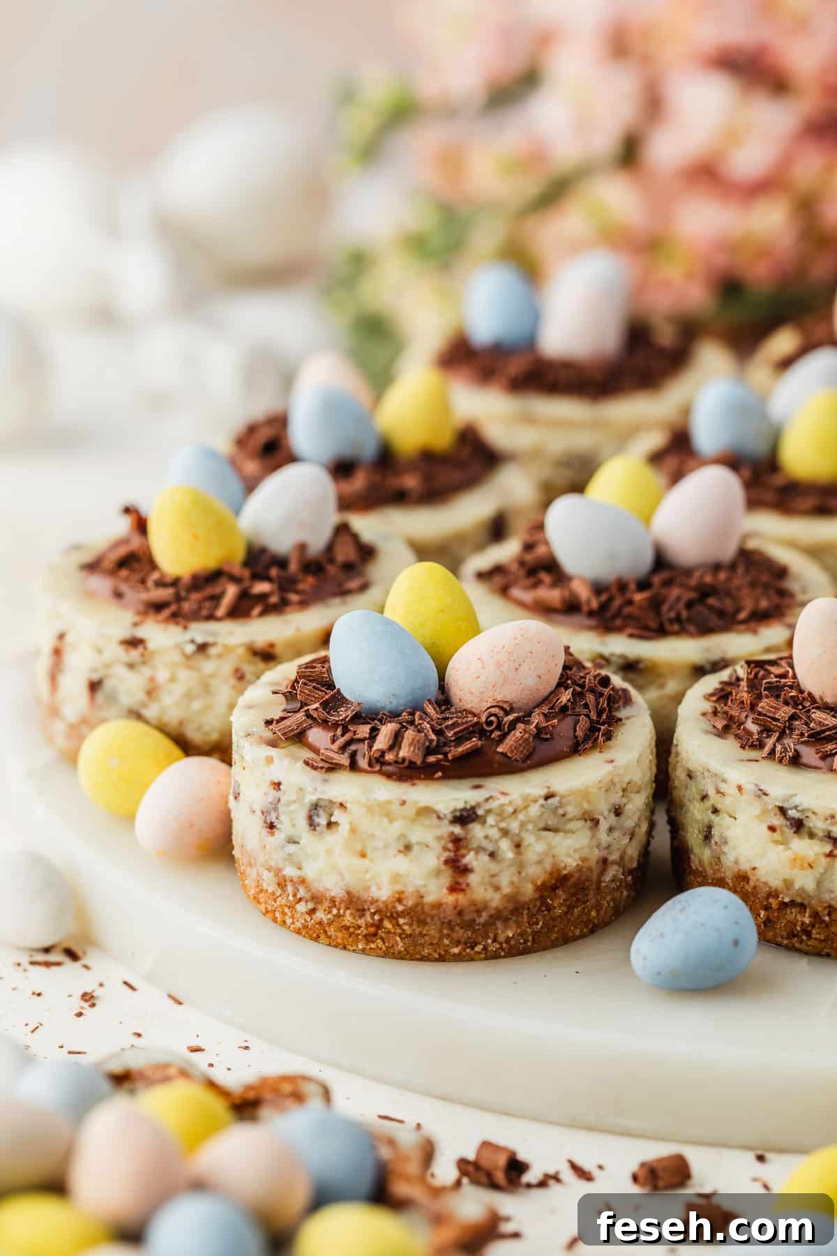 A marble platter of mini egg cheesecakes with ganache nests next to a brown bowl of candies, pink flowers, and white ceramic bunnies on a white counter.