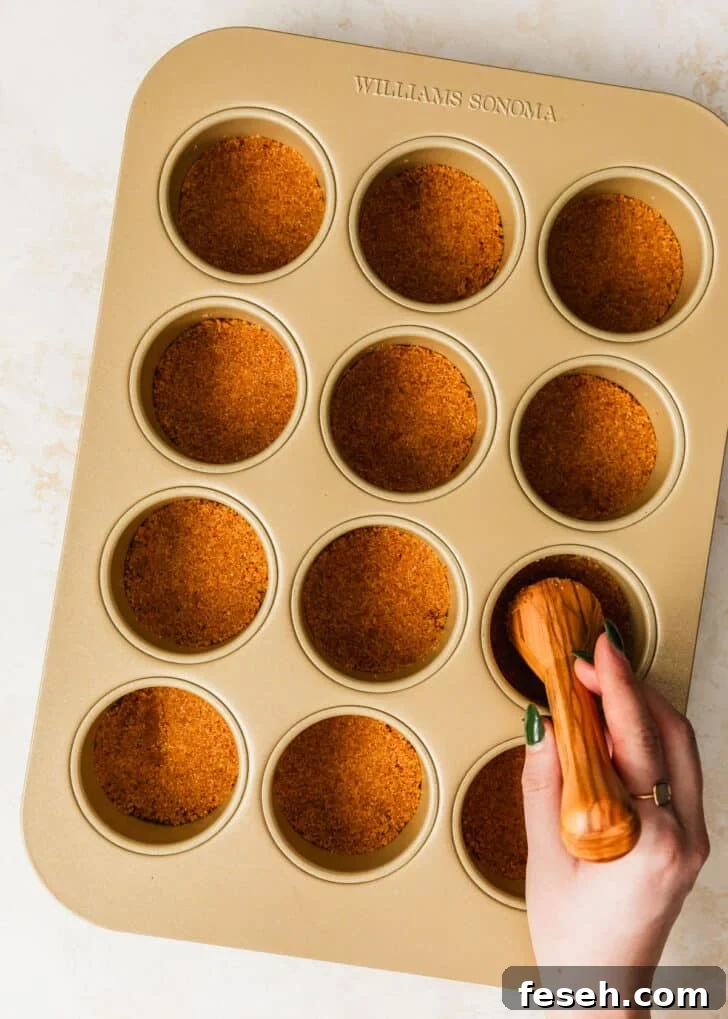 A hand using a pastry tamper to press graham cracker crust into a cupcake pan.