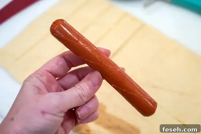 A hand holds a raw hot dog ready to wrap in crescent dough ready to wrap and bake.