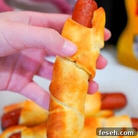 A hand holding an air fryer pigs in a blanket, with more delicious crescent roll-wrapped hot dogs on a plate in the background.