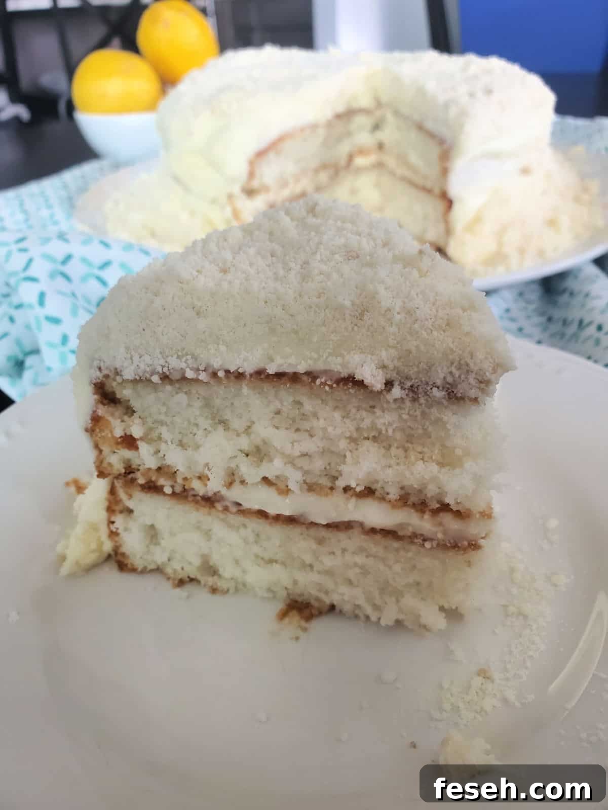 Slice of lemon cake with the rest of the cake behind it next to a bowl of lemons