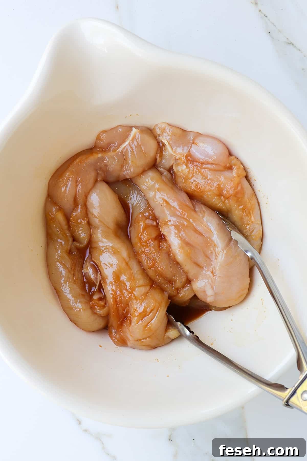Raw chicken tenderloins in a bowl with teriyaki sauce and tongs.