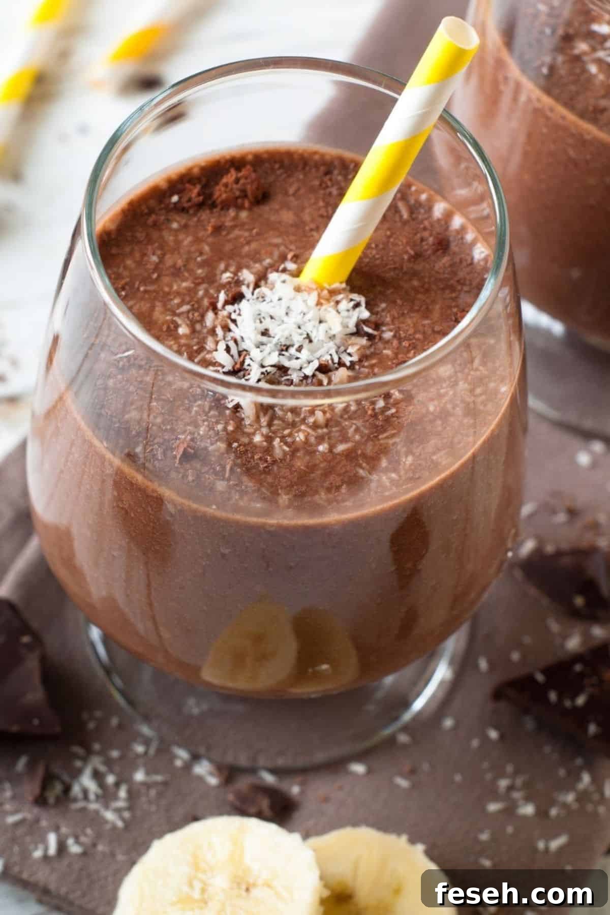 Chocolate Peanut Butter Banana Smoothie Recipe for Creamy Energy 2 A glass of Chocolate Peanut Butter Banana Smoothie topped with shredded coconut, a yellow-and-white striped straw, and banana slices at the base.