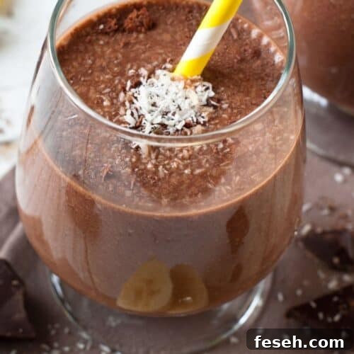 Chocolate Peanut Butter Banana Smoothie Recipe for Creamy Energy 10 A glass of Chocolate Peanut Butter Banana Smoothie with a yellow-and-white striped straw, topped with shredded coconut and surrounded by banana slices and chocolate pieces.