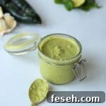 jar of poblano sauce with spoon laying in front and poblano and garlic in background