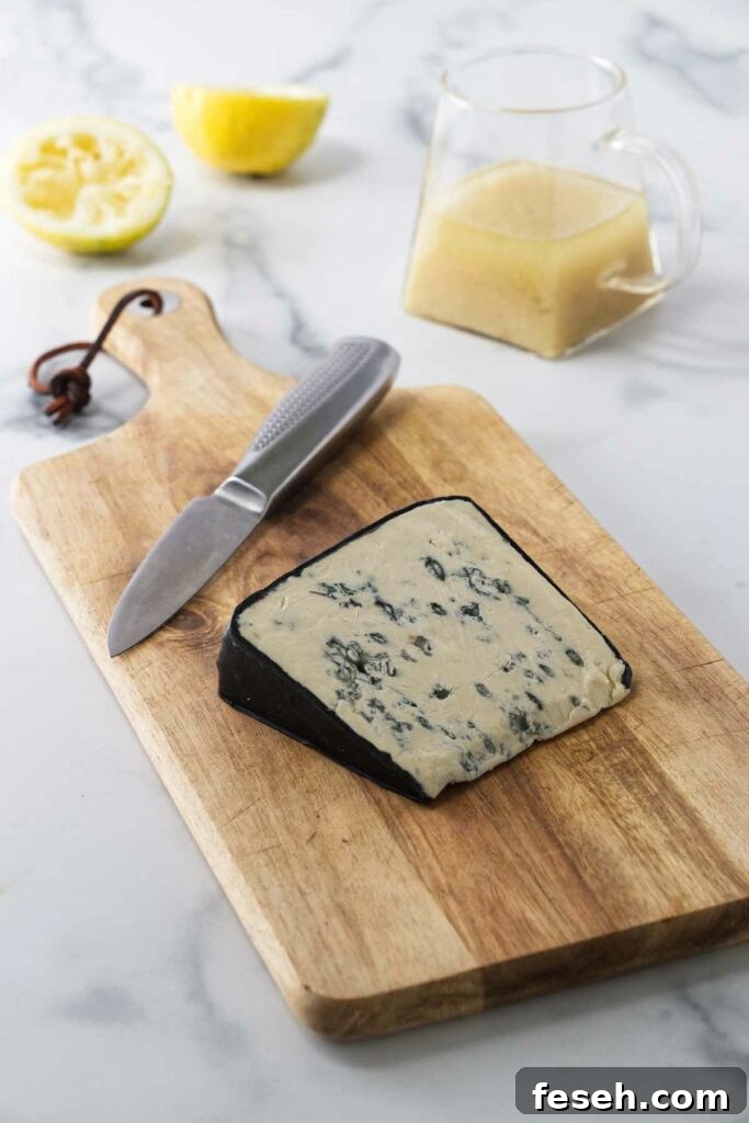 A wooden cutting board with a wedge of blue cheese and a paring knife. A small pitcher of lemon vinaigrette and lemon rinds in the background.