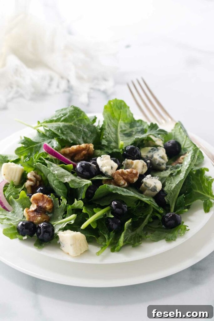 A serving of baby kale and blueberry salad on a salad plate with a fork and a cloth napkin in the background.
