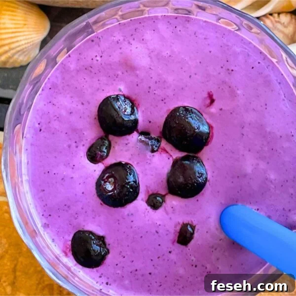 A bluberry banana and peanut butter smoothie with blueberries on top on a tiled background with a blue straw and shells off to the side.