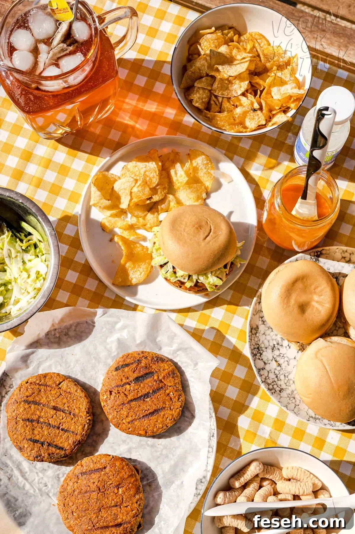 Buffalo Burger on a plate with chips, in a picnic table setting with pitcher of iced tea, plate of burger patties and bowl of chips