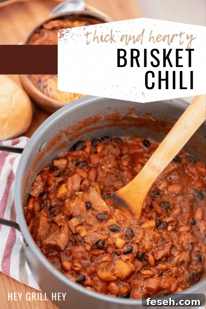 Gray pot full of brisket chili with a bowl of brisket chili and a roll in the background. Text overlay reads: Thick and Hearty Brisket Chili.