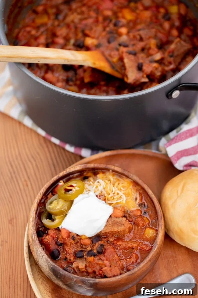 Bowl of brisket chili on a plate next to a roll and metal spoon with a pot of brisket chili in the background.