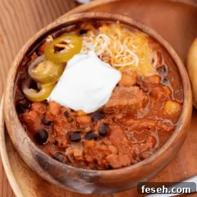 Wooden bowl of brisket chili topped with shredded cheese, sliced peppers, and a dollop of sour cream.