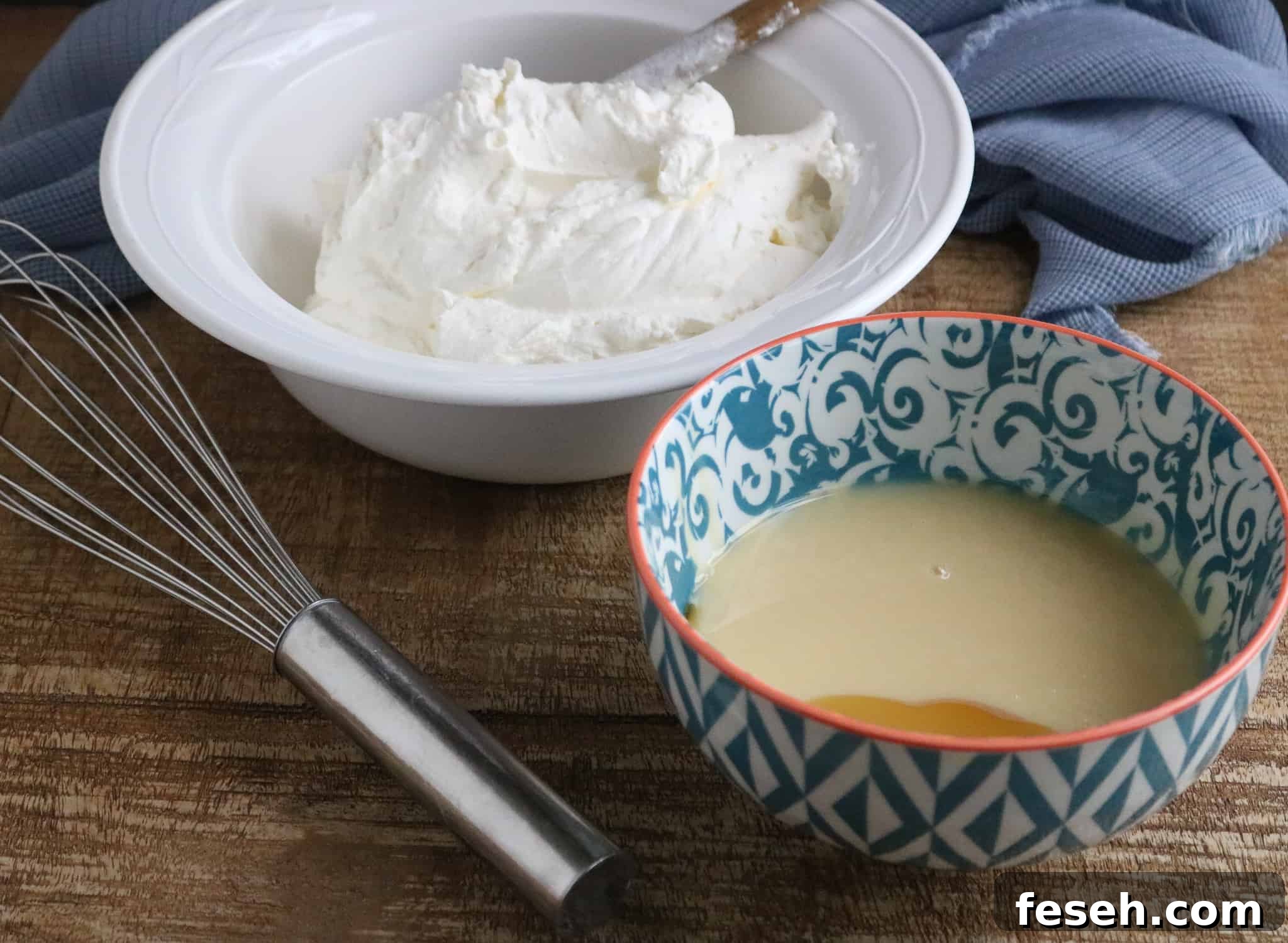 Thick heavy whipping cream in a white bowl and sweetened condensed milk in a teal patterned bowl on a wood table next to a silver whisk and blue kitchen towel